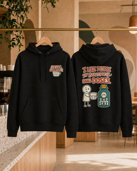 Two black hoodies hanging on wooden hangers on a clothing rack in a cafe setting. The front hoodie features a small graphic on the left chest with the text 'SPECIALIST DOSE' and a coffee cup illustration.