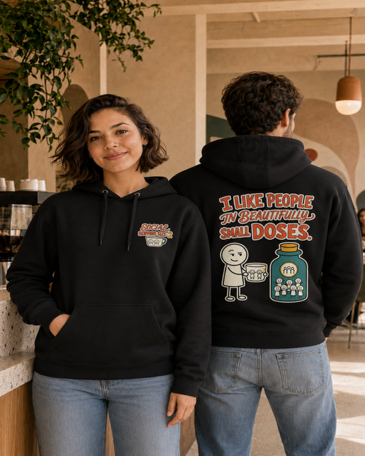 Two models wearing black graphic hoodies in a cafe setting. The woman on the left shows the front of the hoodie with a small chest graphic, and the man on the right shows the back of the hoodie with a large cartoon graphic and text reading 'I LIKE PEOPLE IN BEAUTIFUL SMALL DOSES.